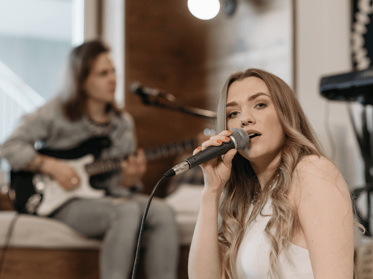 two women in a studio, one holding a microphone singing, the other in the background is playing guitar